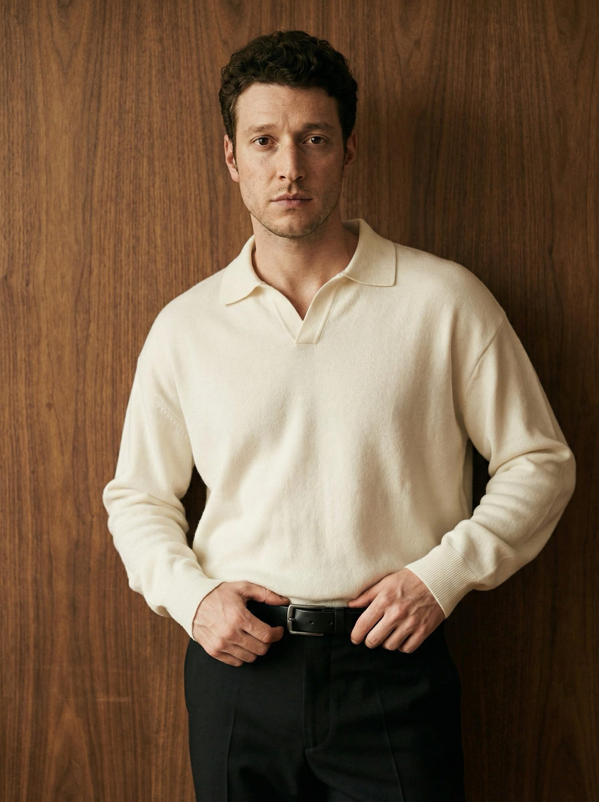 Portrait of a man in an ivory turtleneck sweater against a warm walnut wood panel, hands on waist, confident expression.
