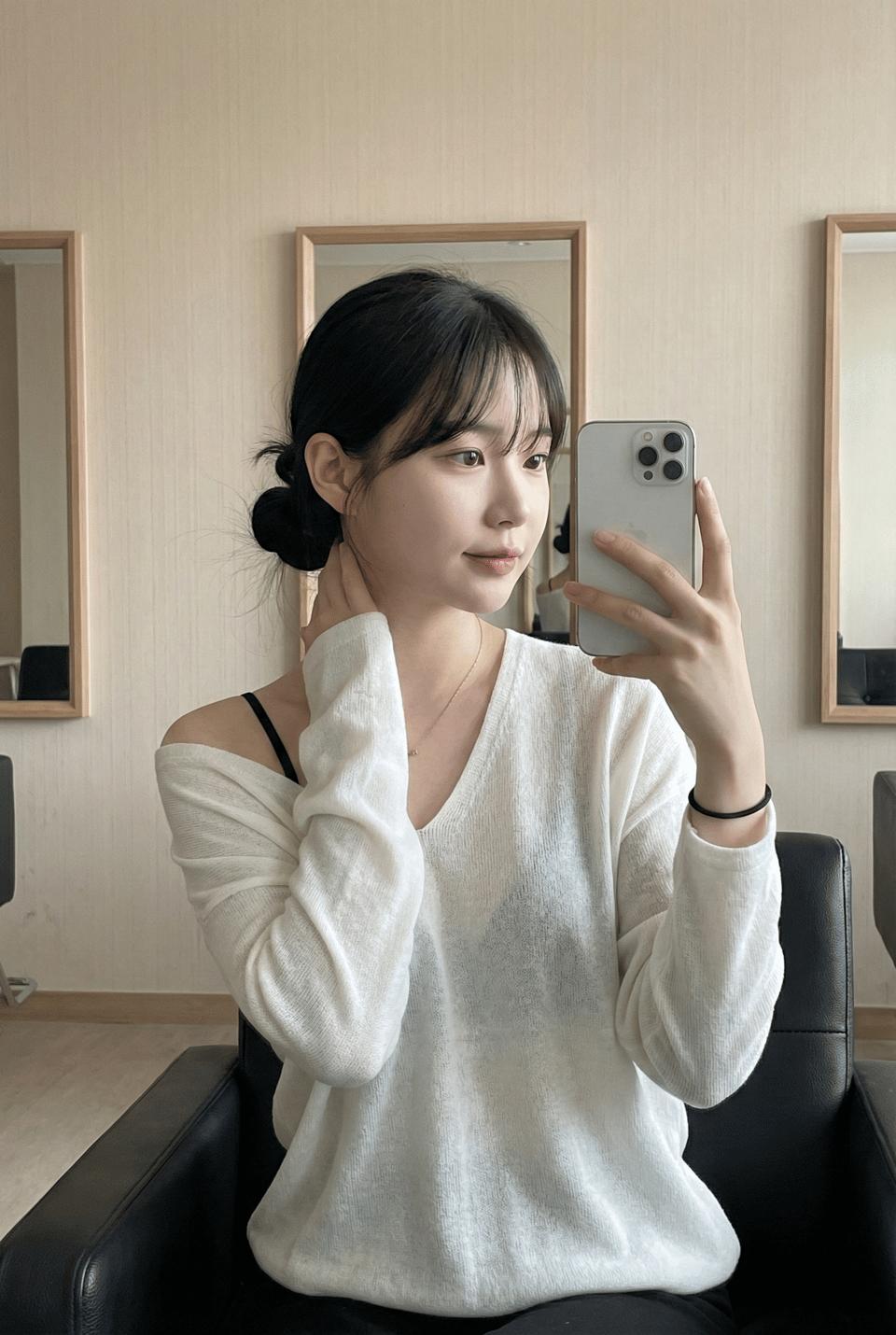 A serene Asian woman in a mirror selfie, wearing a deep V-neck top with hair tied back, relaxing on a leather chair.