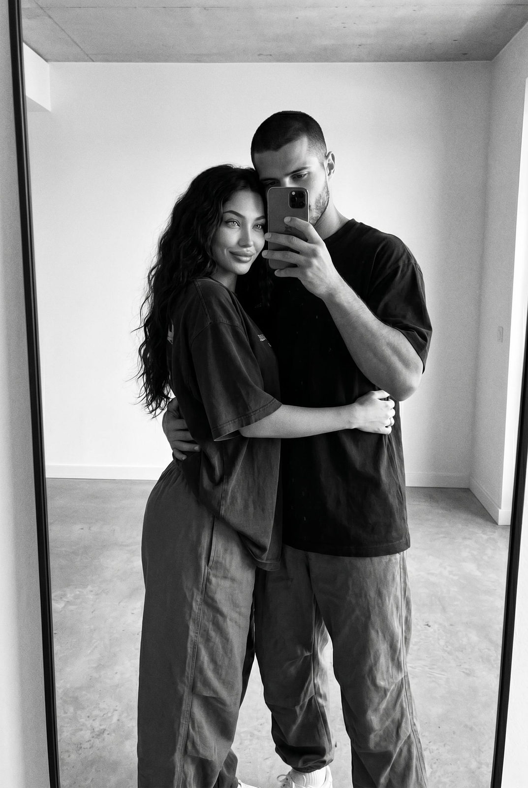 A stylish couple taking a black and white mirror selfie in a modern apartment with neutral walls and soft lighting.