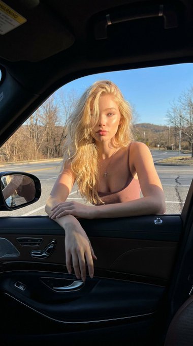 A woman leans through an open car window, resting her forearms on the door frame, with long wavy hair and a dusty rose top.