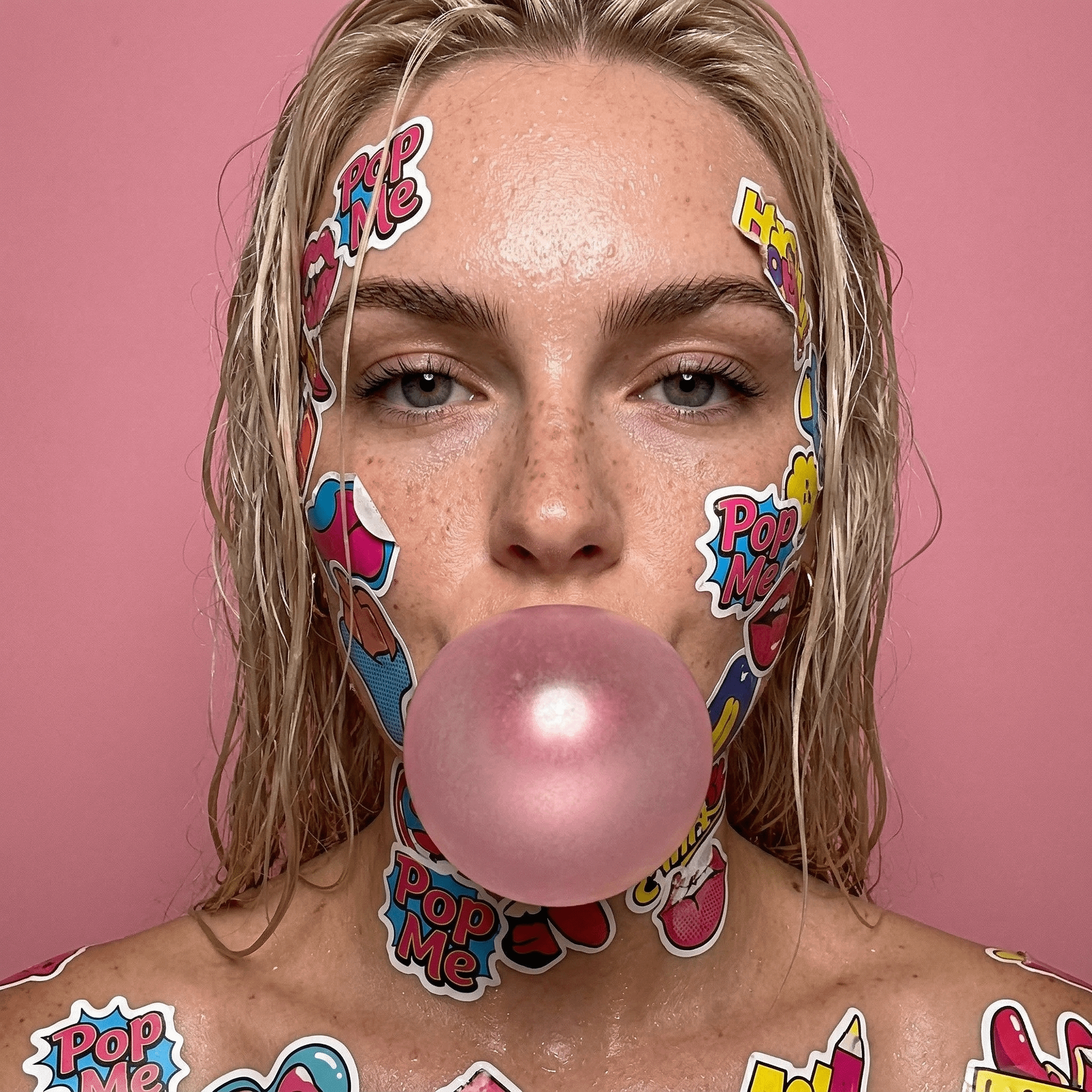 Close-up portrait of a person blowing a large pink bubblegum bubble with colorful comic book-style stickers on their skin.
