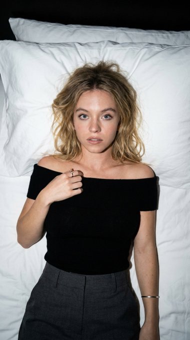 Portrait of a woman lying on a white bed, wearing a black off-the-shoulder top and gray trousers, with distinct shadows.