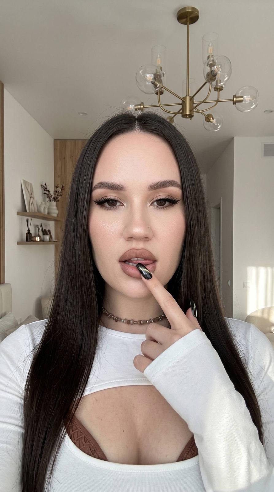 A girl with long straight hair gazes into the camera, bright modern room backdrop with chandelier, morning light accentuating her features.
