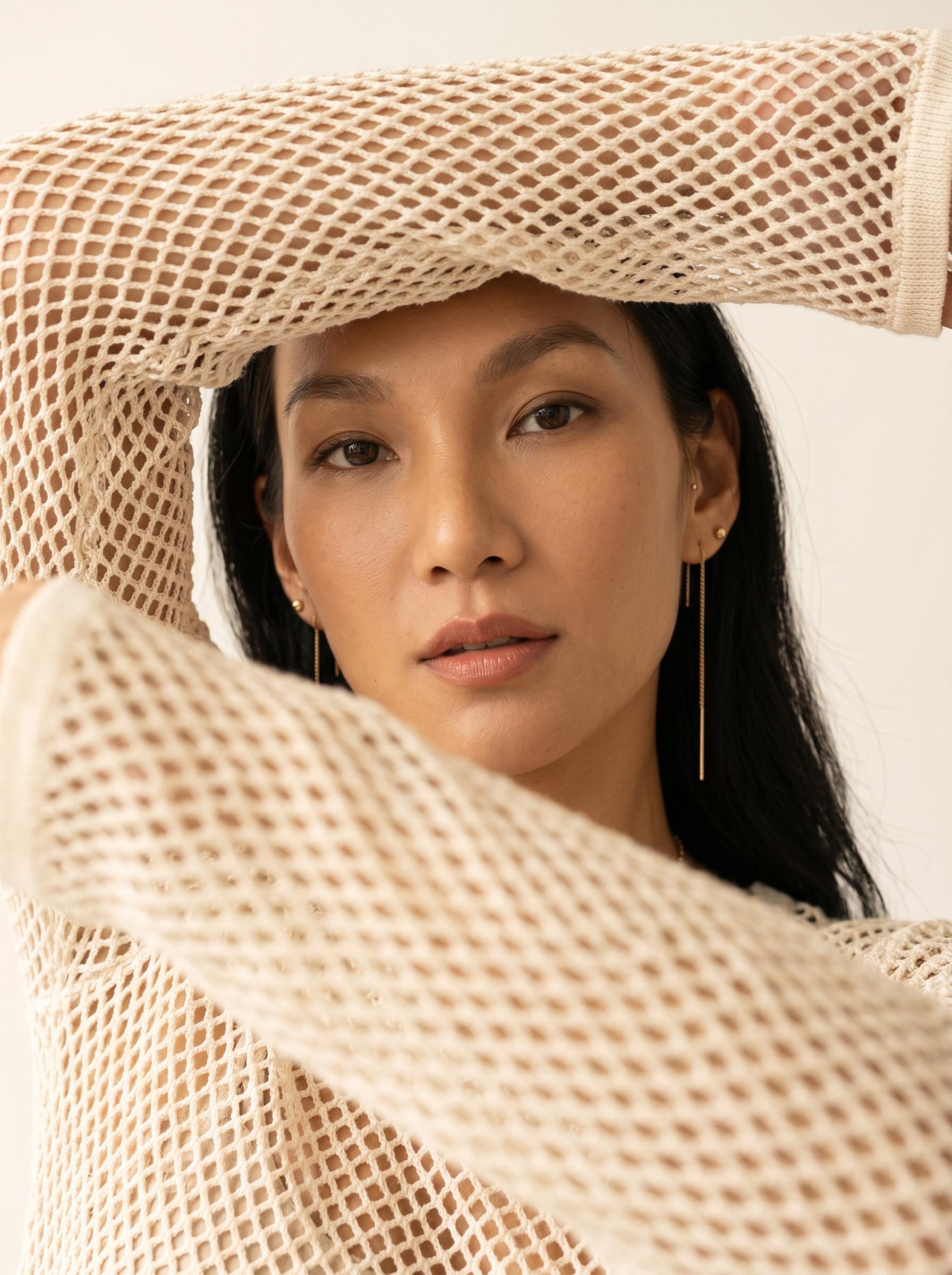A person in a cream-colored mesh sweater with arms raised, framing their serene face, background softly blurred.