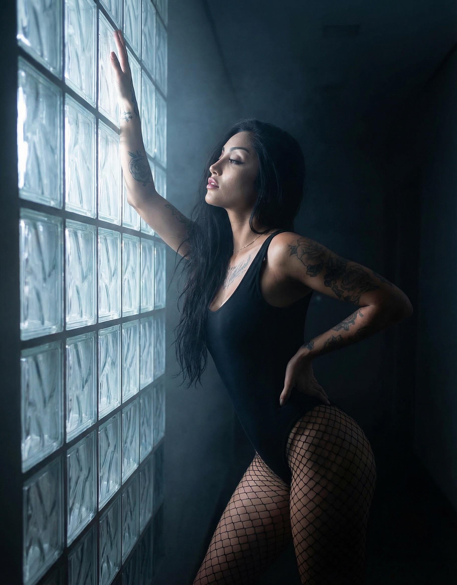 A woman in a black bodysuit gracefully poses in a glass brick corridor bathed in moonlight.