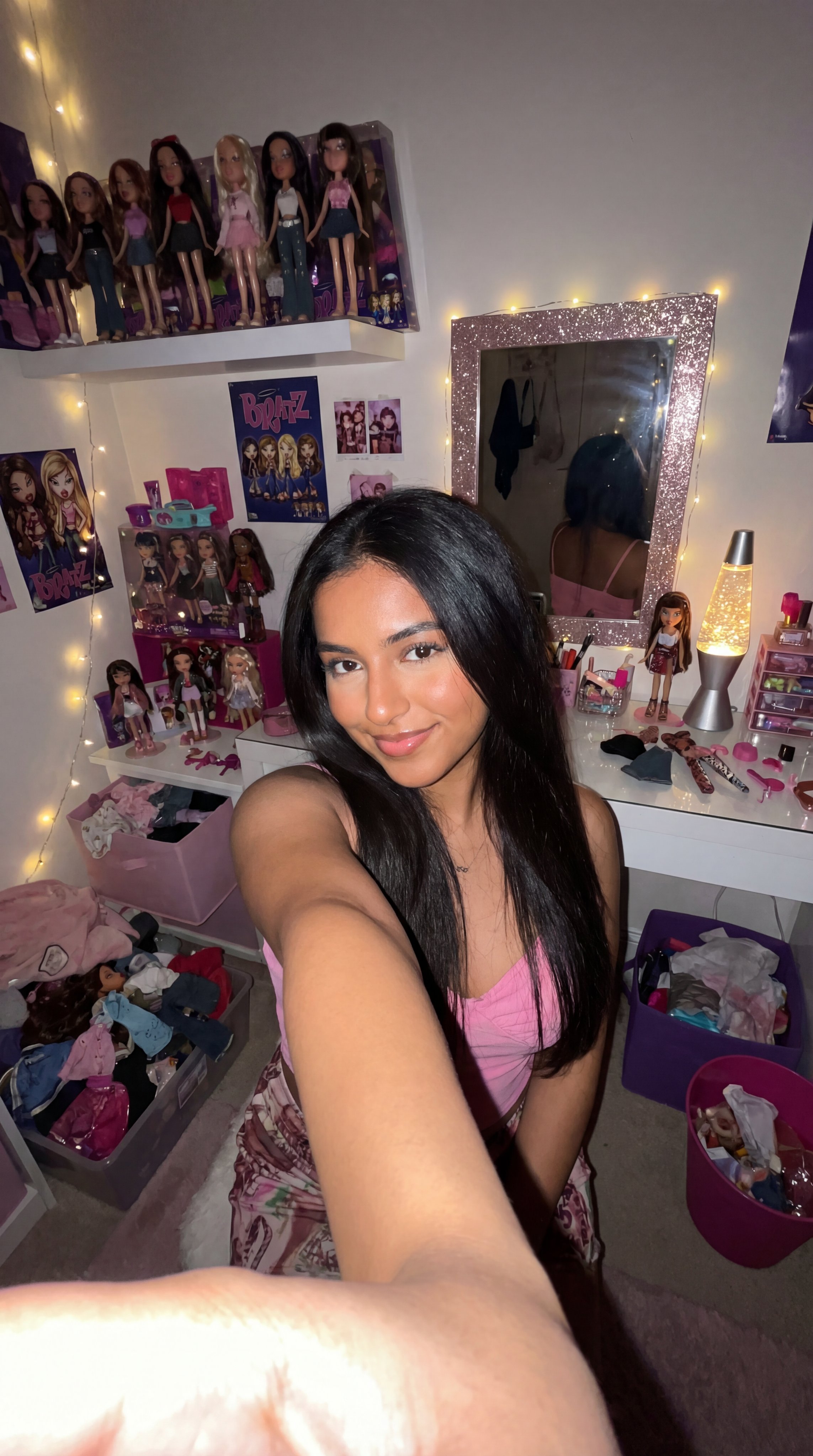 Stylish young woman with a warm tan skin tone in a pink bedroom full of Bratz dolls and glittery decor, posing with a charming smile.
