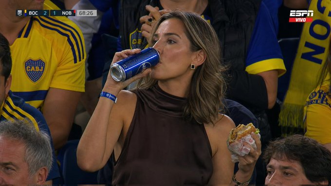 A woman in a brown satin halter top sits in a stadium, holding a burger and a blue can, wearing gold earrings.