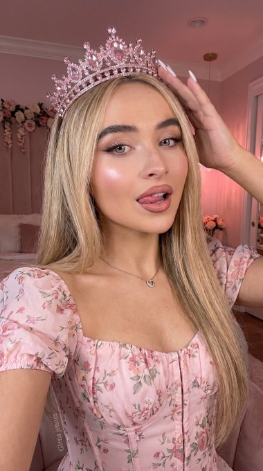 A close-up selfie of a person wearing a sparkling pink tiara, light pink corset, and a playful expression in a soft pink bedroom.