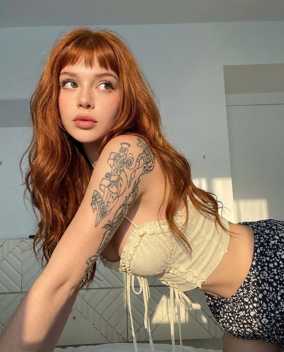 A woman with long wavy copper-red hair and a floral skirt in a sunlit bedroom, featuring warm light and intricate tattoos.