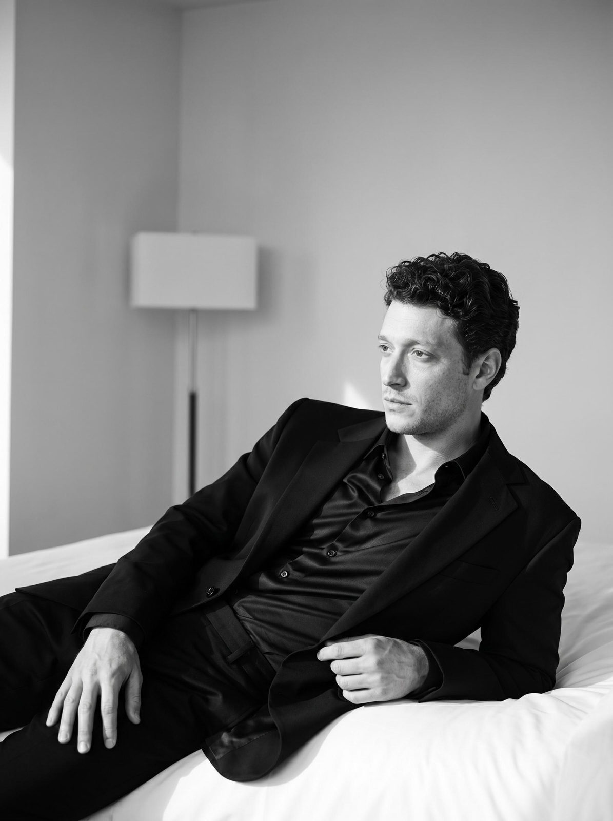 A man in a loose-fitting black suit reclines on a hotel bed, showcasing dramatic lighting and soft shadows.
