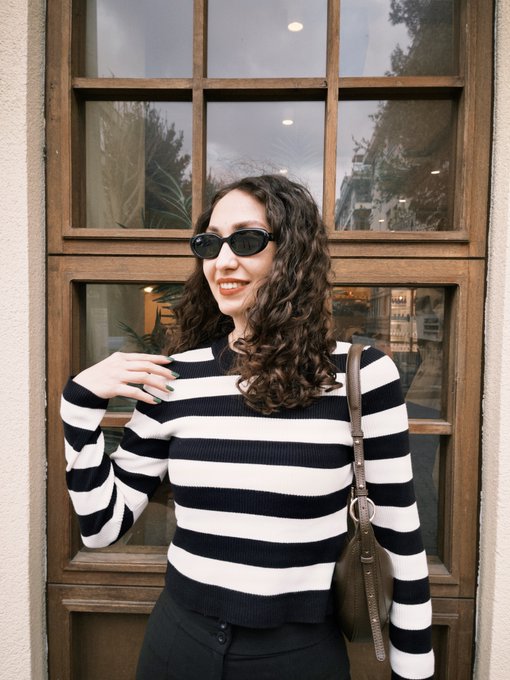 A person in a long-sleeved ribbed knit sweater poses at an eye-level angle, showcasing a large wooden window and soft natural light.