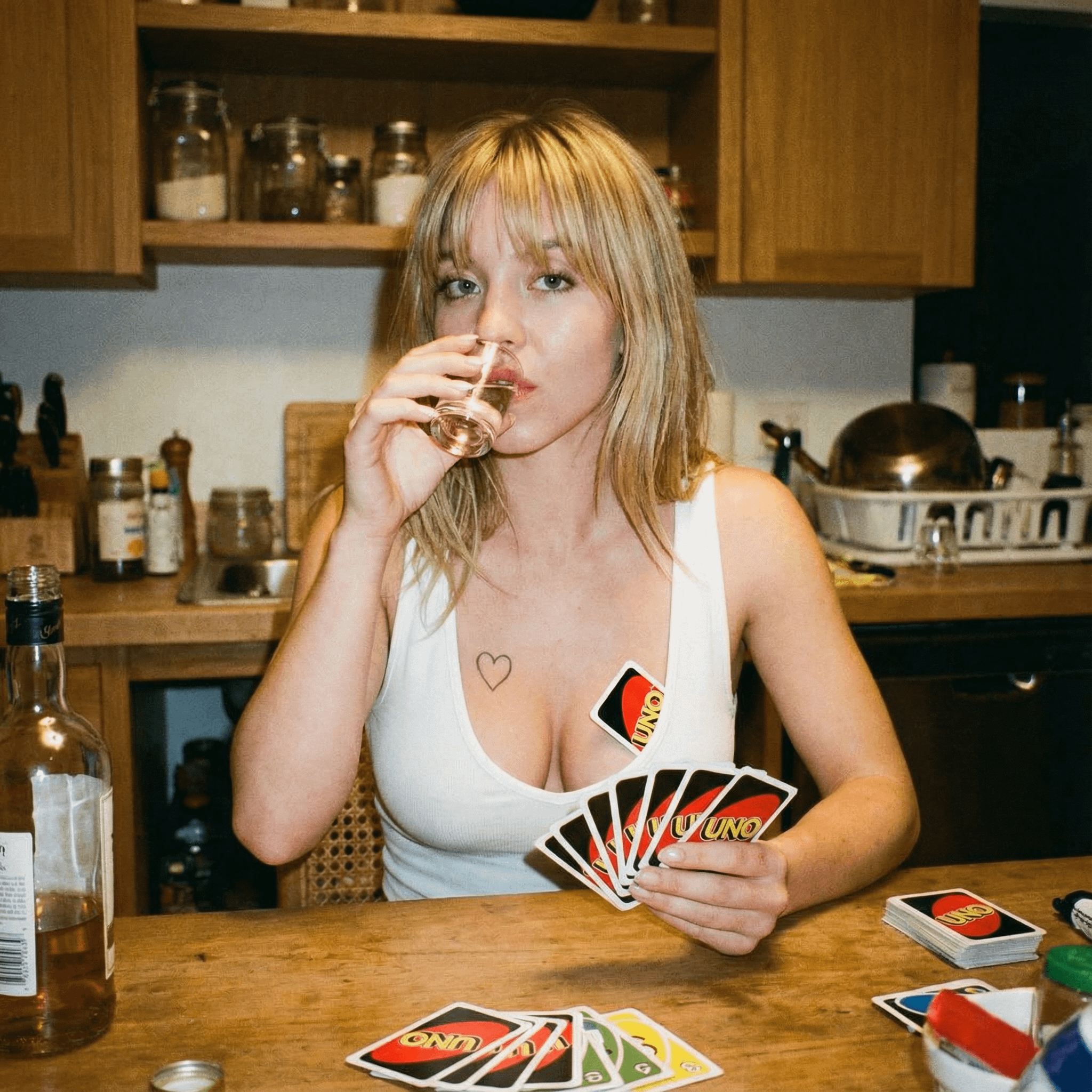 A woman sitting at a wooden kitchen table, holding a fan of UNO cards while drinking from a shot glass, warm lighting enhances the scene.