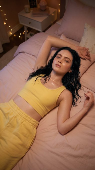 Overhead view of a person on a pink bed with dark hair spread out, wearing a yellow tank top and sweatpants in warm light.