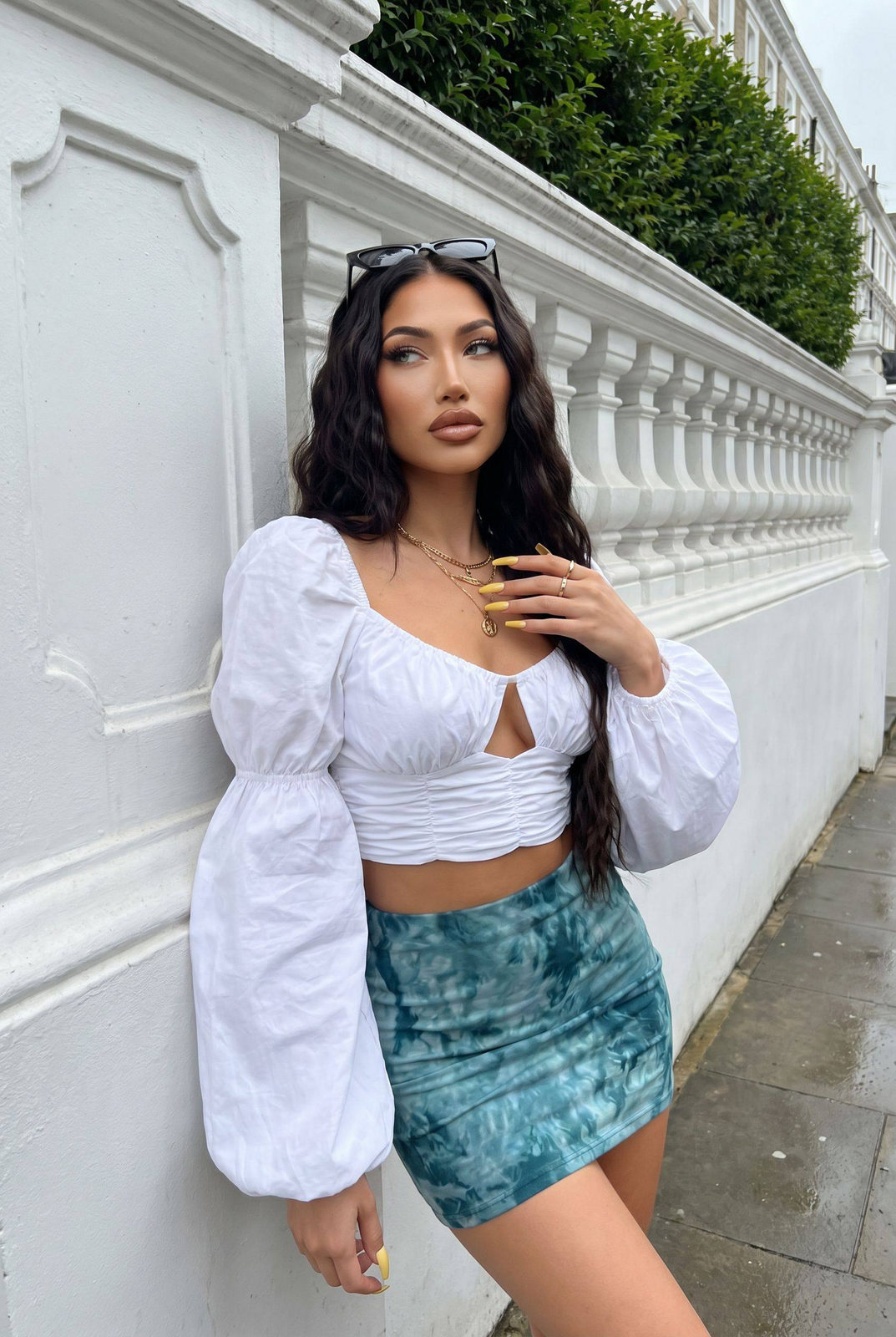 A woman leans against a large ornate white wall wearing a stylish white crop top and a tropical tie-dye mini skirt.