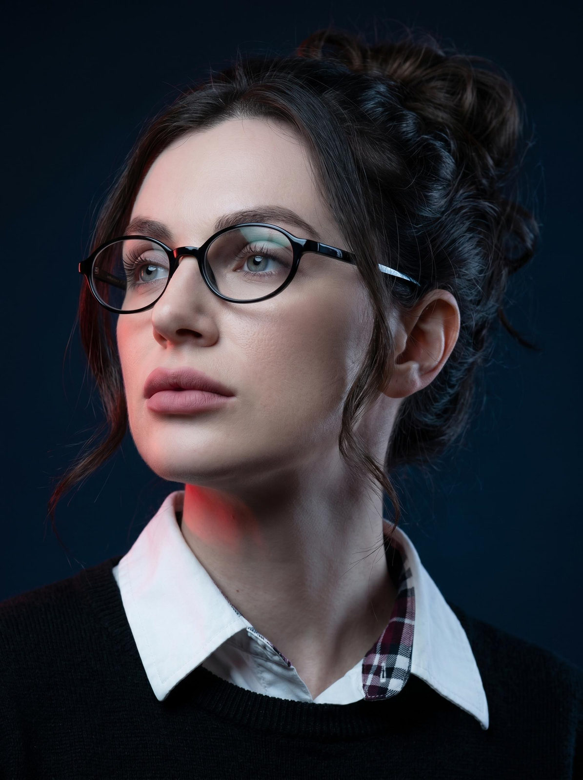 Close-up portrait of a woman with wavy bun, neutral cool makeup, wearing glasses, set against a dark blue-black background.