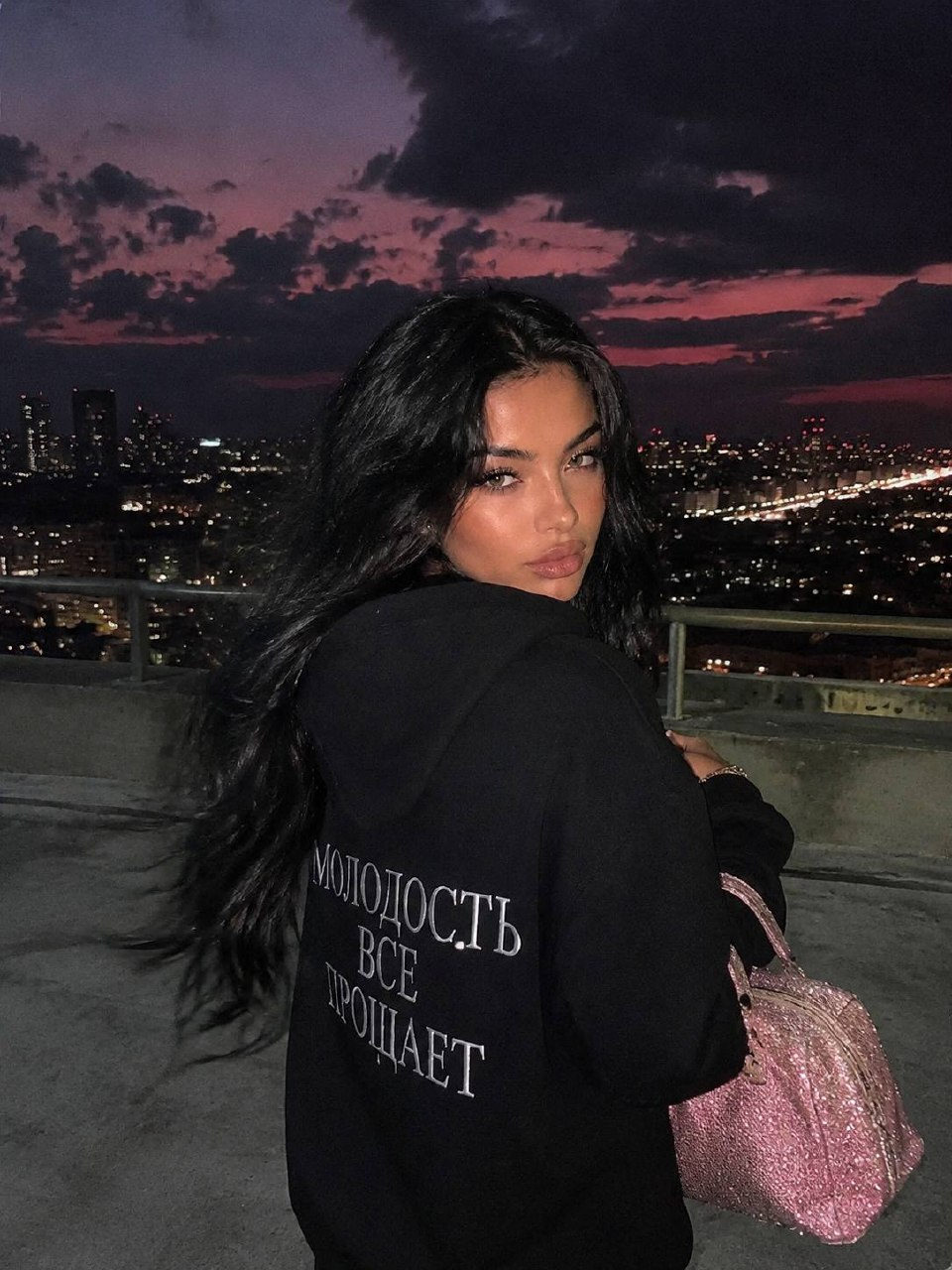 Ultra-realistic Y2K rooftop portrait of a woman in a black hoodie, gazing calmly at the camera, with a pink glitter bag.