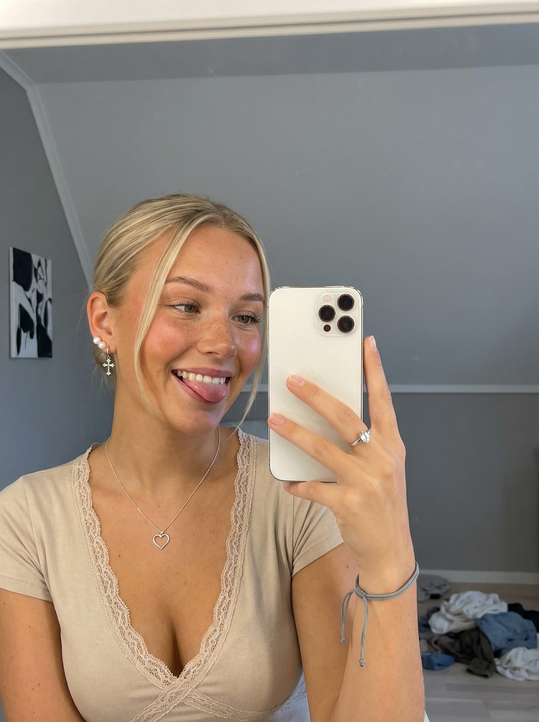 A woman taking a mirror selfie with warm undertones, freckles, and blue eyes, smiling playfully in natural light.