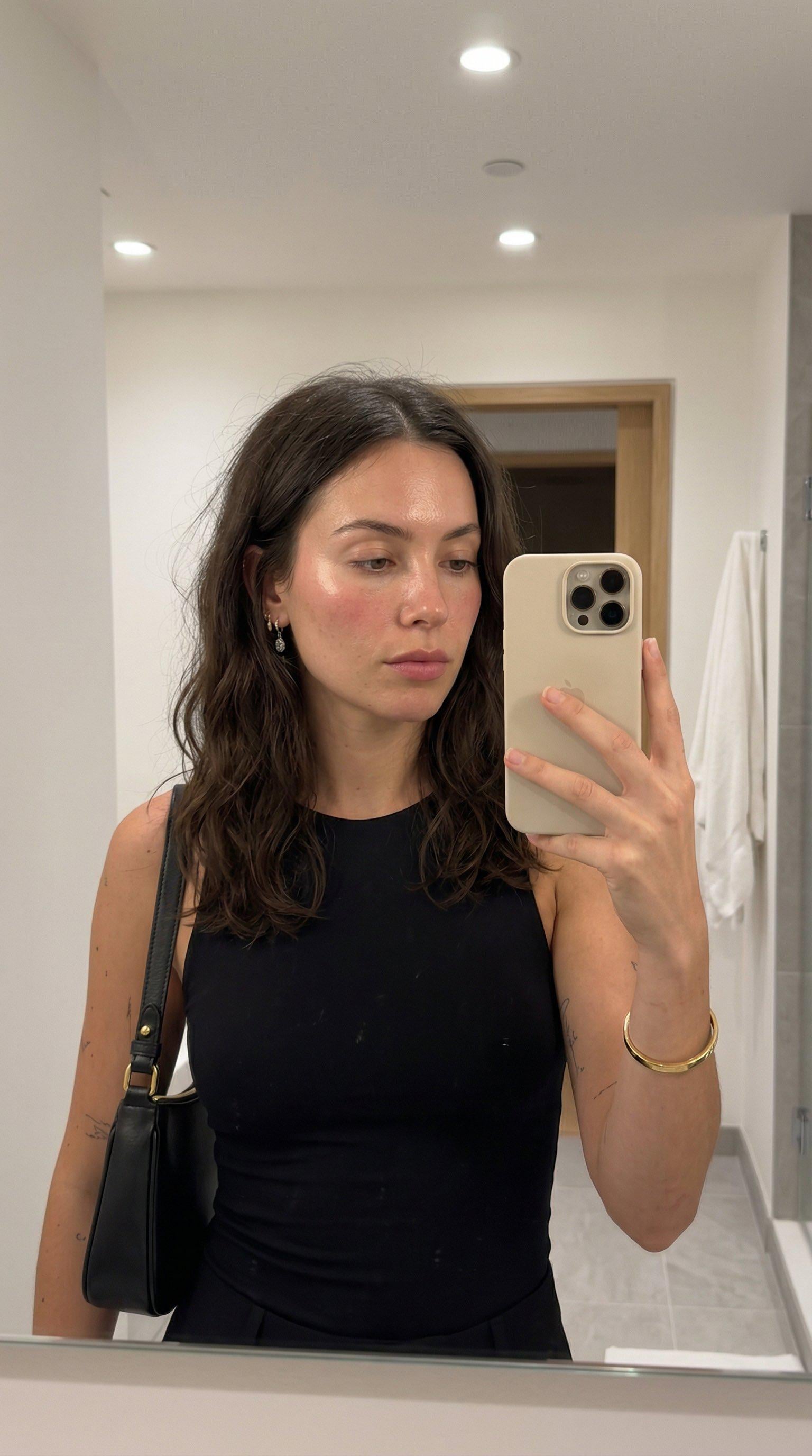 A woman in a modern bathroom holding a beige smartphone, showing neutral expression while looking at her reflection.