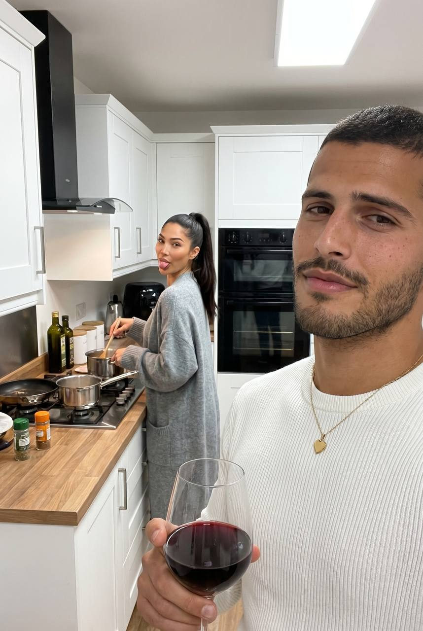 A man in a white sweater takes a selfie in a bright kitchen with a woman cooking in the background.