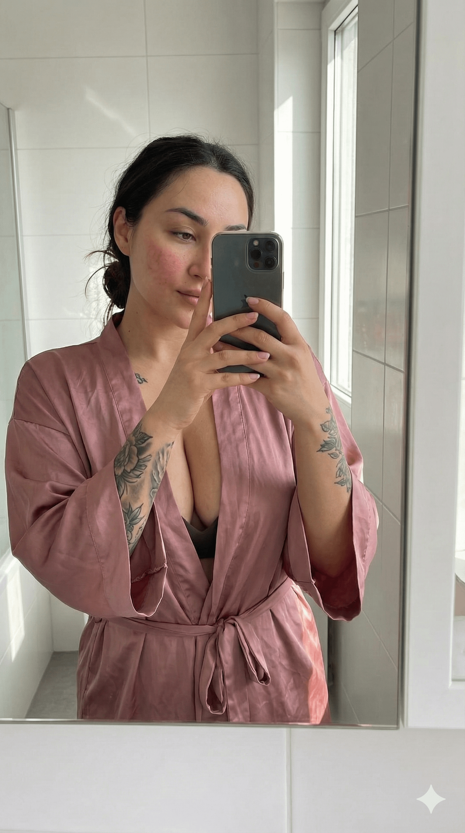 A woman in a soft pink silk robe standing in a bright bathroom, casually holding a smartphone and caught mid-gesture in a natural pose.