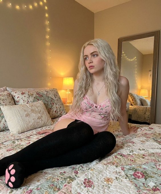 Woman in a pink lace camisole with floral quilt background, sitting on a bed with black thigh-high socks and warm ambient lighting.