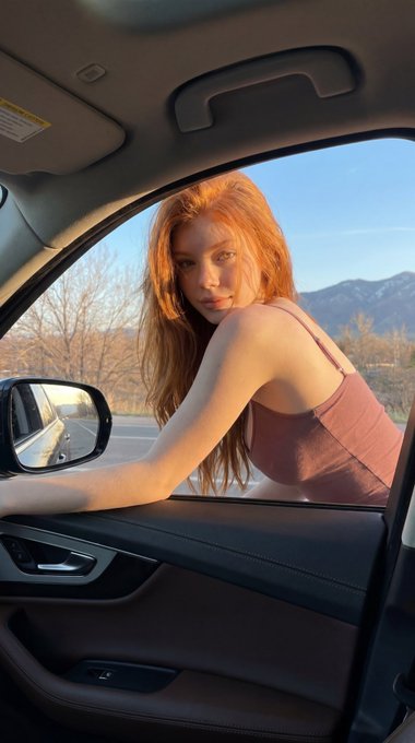 A young woman with long wavy red hair leans through an open car window, wearing a mauve tank top, in a barren landscape during golden hour.