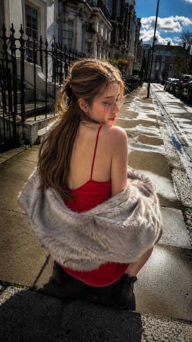 A woman in a bright red slip dress crouches on a stone step, draped in a light grey faux fur stole, gazing over her shoulder.