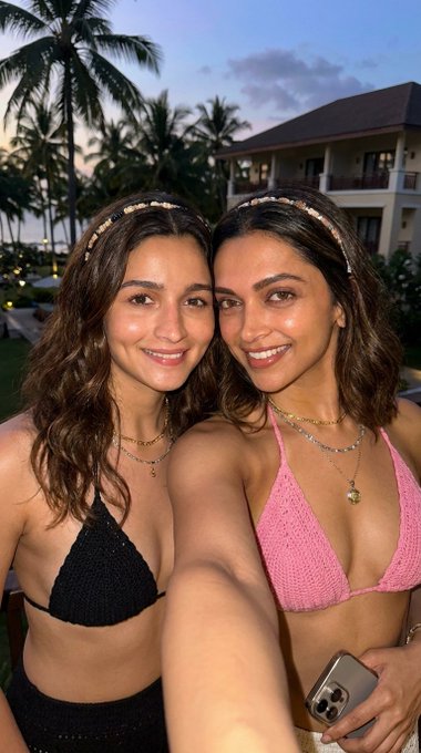 A cheerful young Indian woman in a black crocheted bikini takes a selfie at a tropical resort during golden-hour, with palm trees in the background.