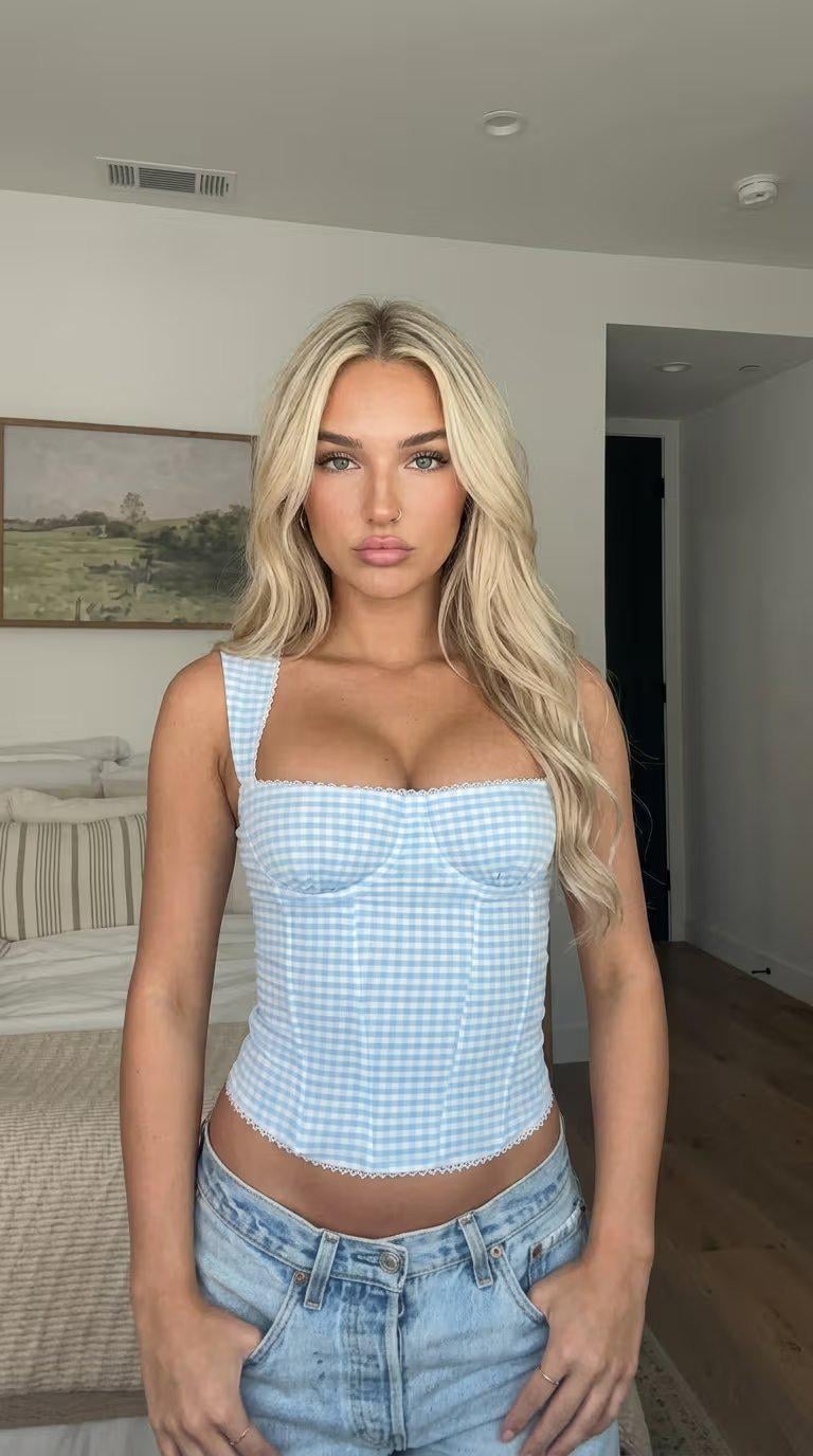 Woman in a light blue and white gingham corset top, standing in a bright bedroom with natural light.