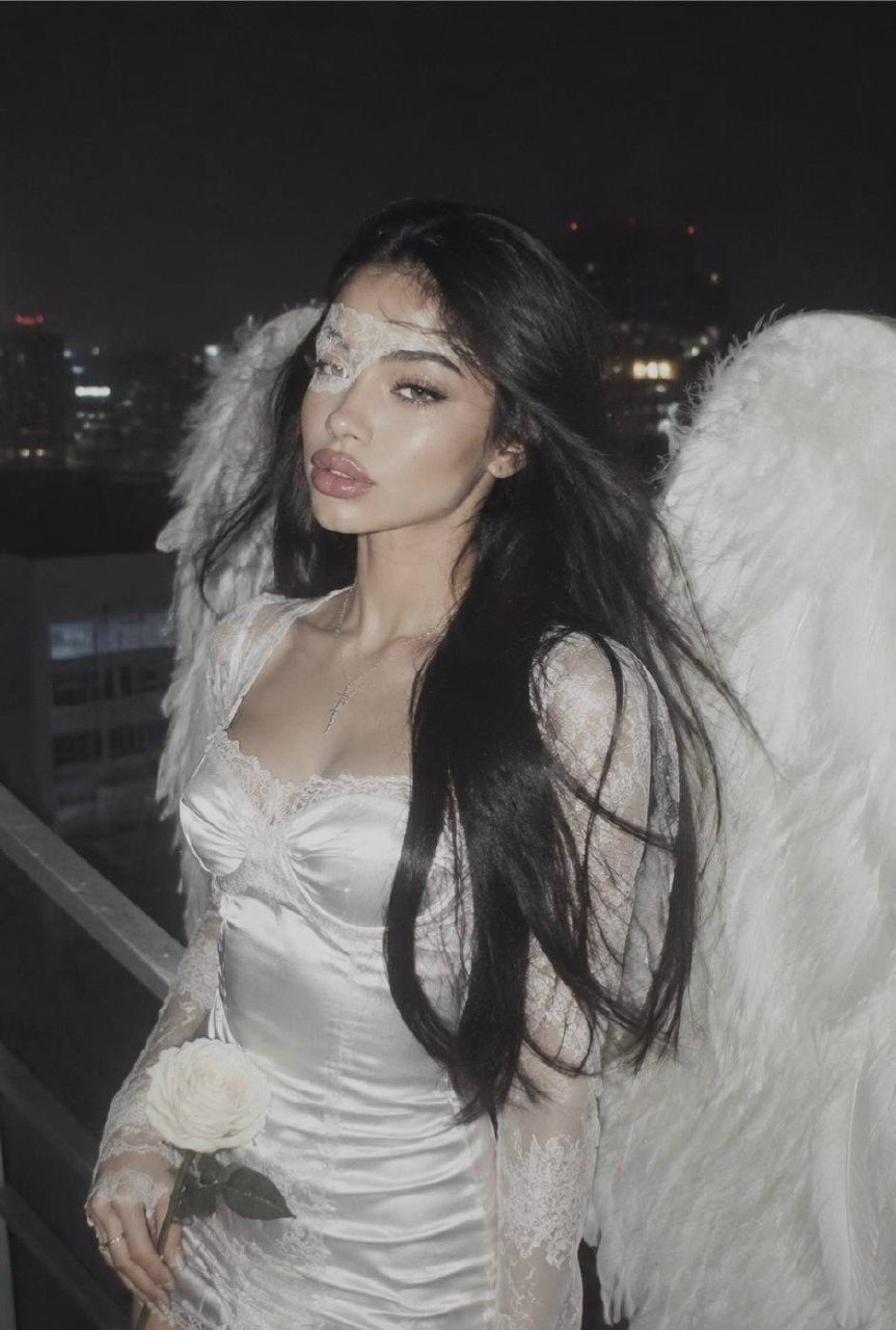 A woman in a fitted white satin mini dress stands on a rooftop at night, large angel wings spread behind, holding a white rose.