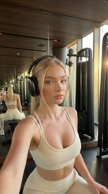 A woman takes a selfie in a gym wearing a beige sports bra and high-waisted leggings, with golden sunlight streaming in.