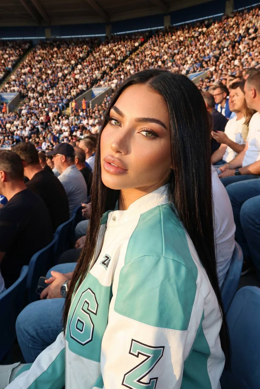 Woman in a mint racing jacket seated at a crowded stadium, looking back with a sultry gaze against warm golden hour light.