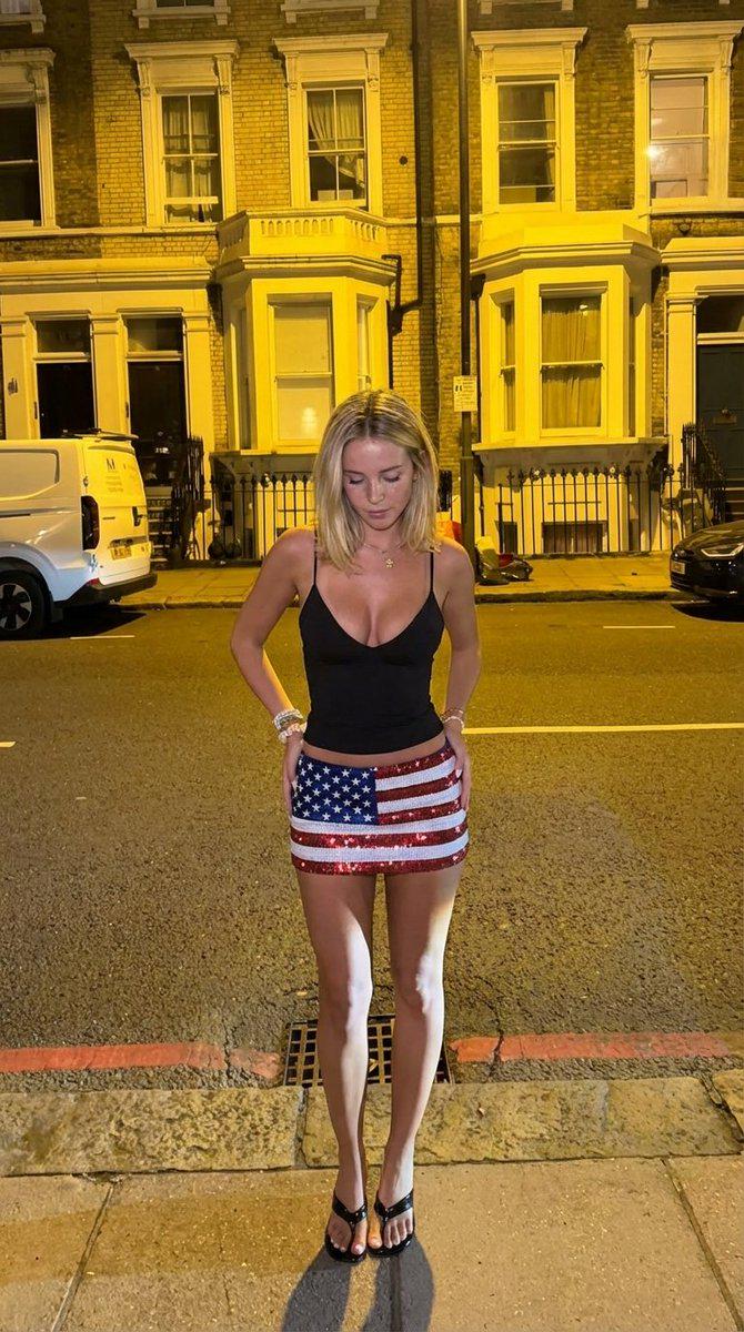 A woman in a black spaghetti strap tank top and an american flag sequined skirt stands on a night street, illuminated by warm street lights.