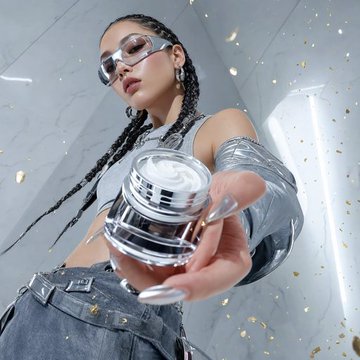 A model holding a circular cosmetic jar with white cream, adorned in futuristic silver sunglasses and grey attire, against a marble background.