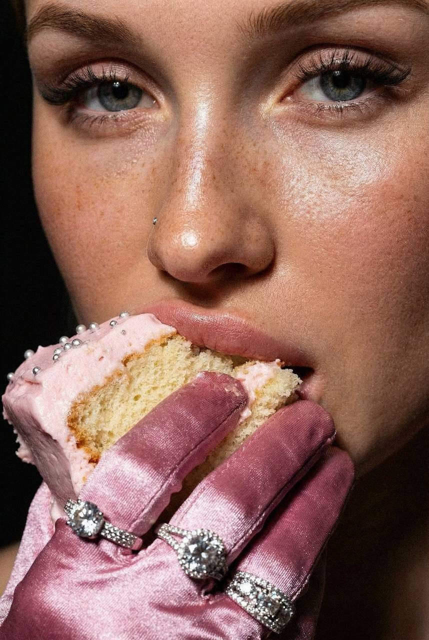 Extreme close-up of a woman's face holding a partially bitten vanilla cake with a satin pink glove.