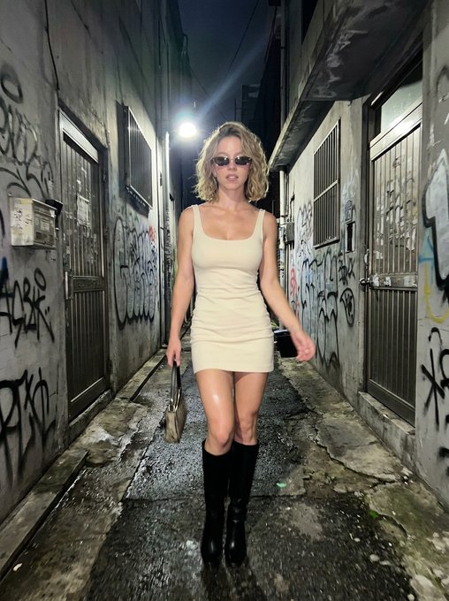 A confident woman in a low-neck fitted mini dress walking through a narrow urban alley at night, grungy graffiti walls and dim warm lighting around her.