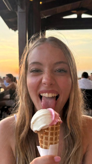 Close-up of a woman holding a waffle cone with vanilla and strawberry swirl ice cream, warm evening light in the background.