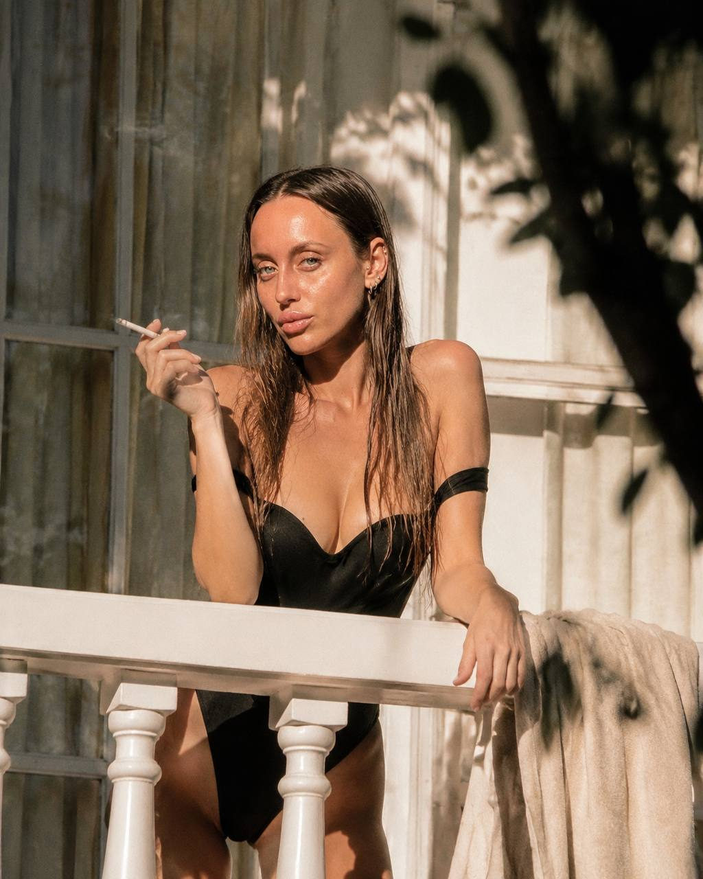 Portrait of a woman in a black swimsuit on a white balcony, relaxed pose with a cigarette.