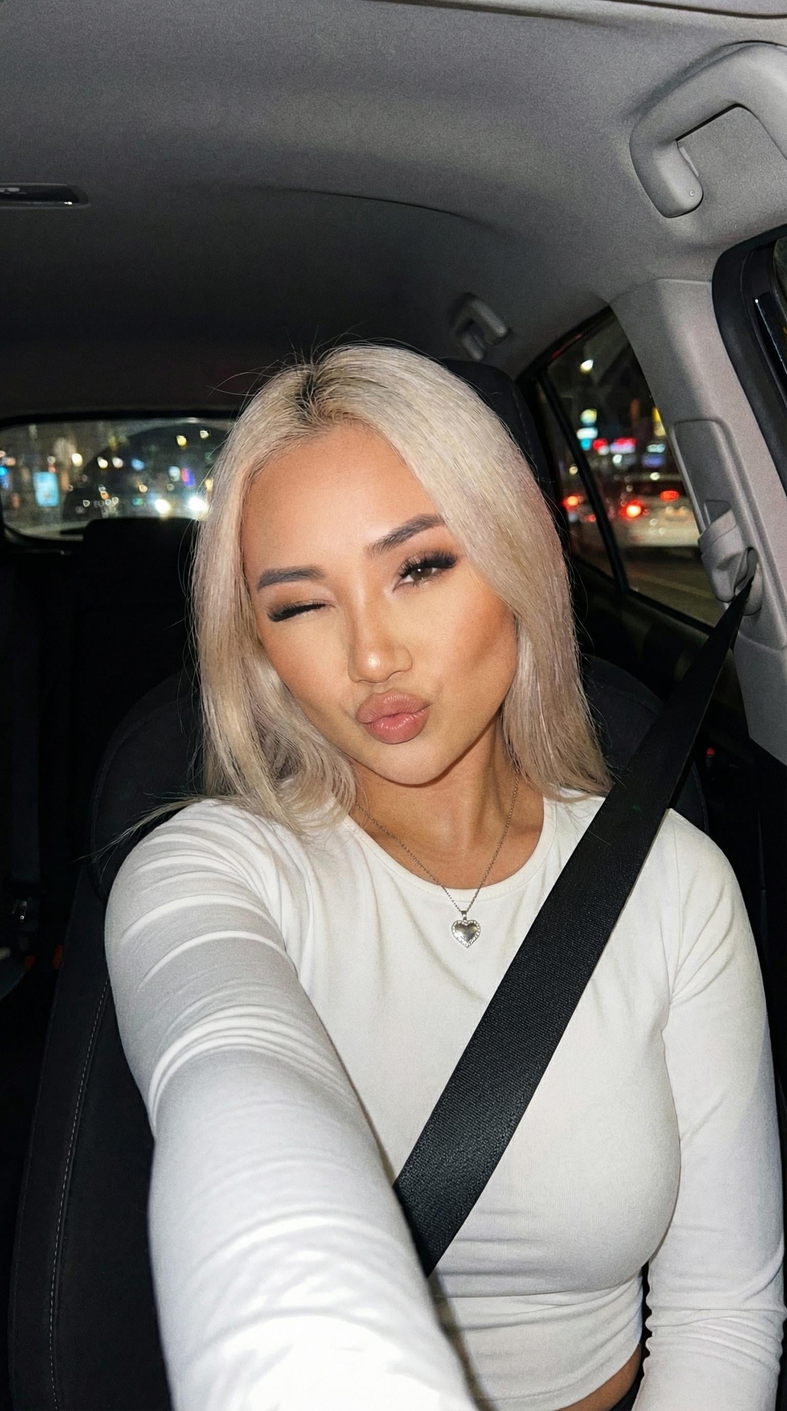 A playful selfie featuring a person wearing a heart-shaped necklace, taken inside a car at night.