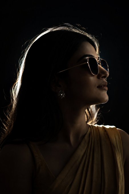 A woman in gold-rimmed aviator sunglasses featuring dark lenses, illuminated by strong backlight that creates a halo effect, wearing a textured mustard-yellow garment.