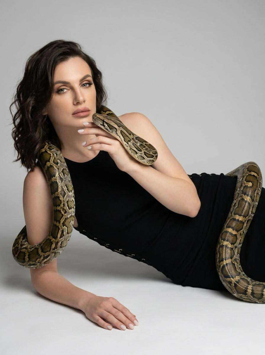 A woman lies on the studio floor, supported by one arm, with a large python coiled around her shoulders and legs, gazing past the camera.