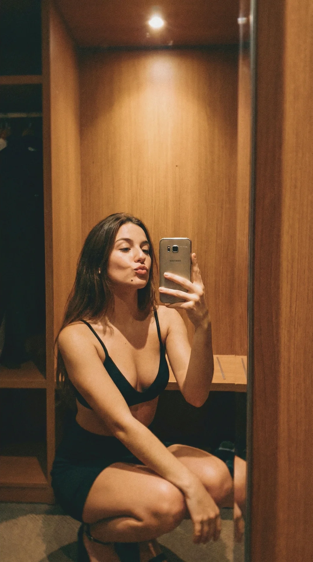 A young woman with long dark brown hair posing in front of a narrow hotel closet mirror, wearing a black bralette and mini skirt, taking a selfie.