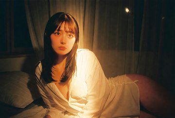 A woman reclined by a bedroom window, wearing a white longline shirt, warm indoor lighting accentuating fabric textures.
