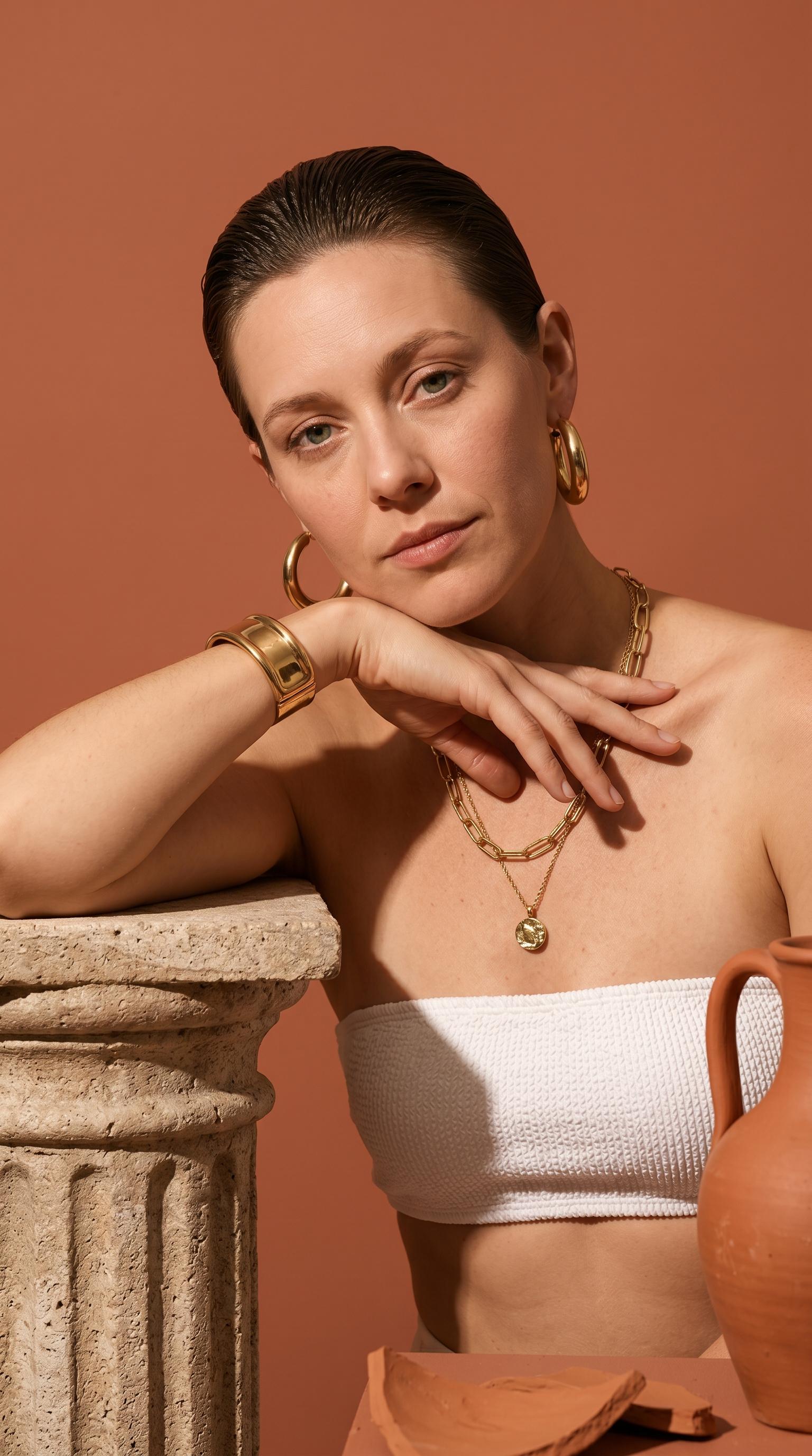 A woman in a white bandeau top adorned with gold jewelry, standing beside a textured stone column in a studio.