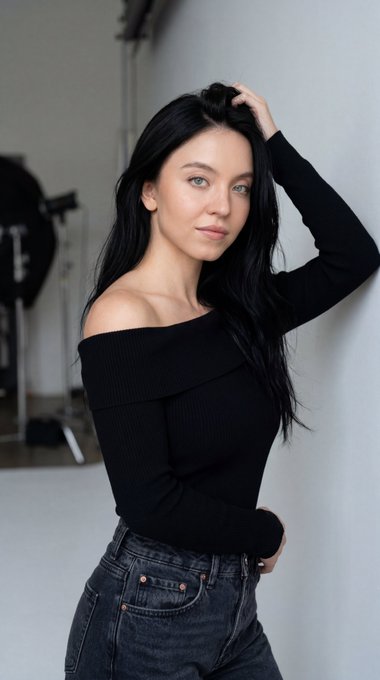 A woman with long dark hair leans against a plain white wall in a professional studio, wearing a black ribbed knit top and dark jeans.