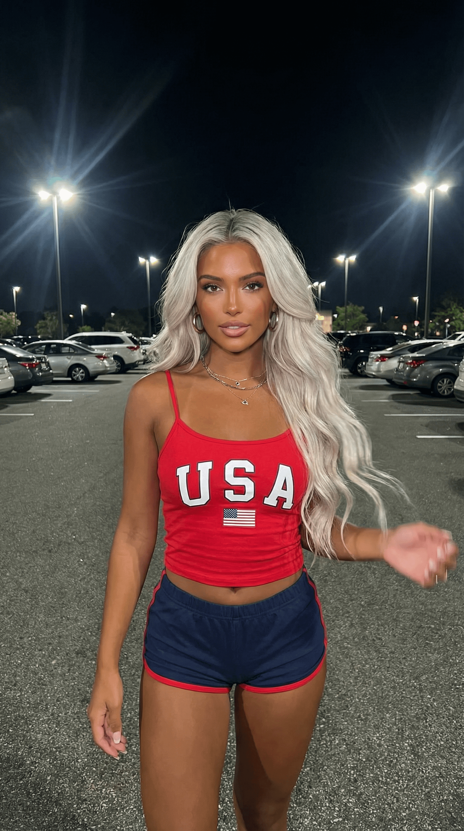 A woman in a red crop top and navy athletic shorts standing in an outdoor parking lot at night, illuminated by streetlights.