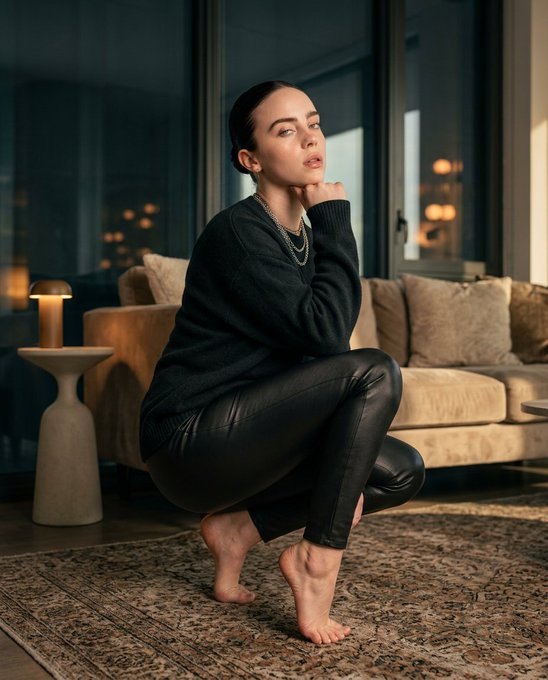 A woman crouching on a vintage Persian rug in a dim living room, wearing a dark knit sweater and leather trousers, with a soft glow from a lamp.