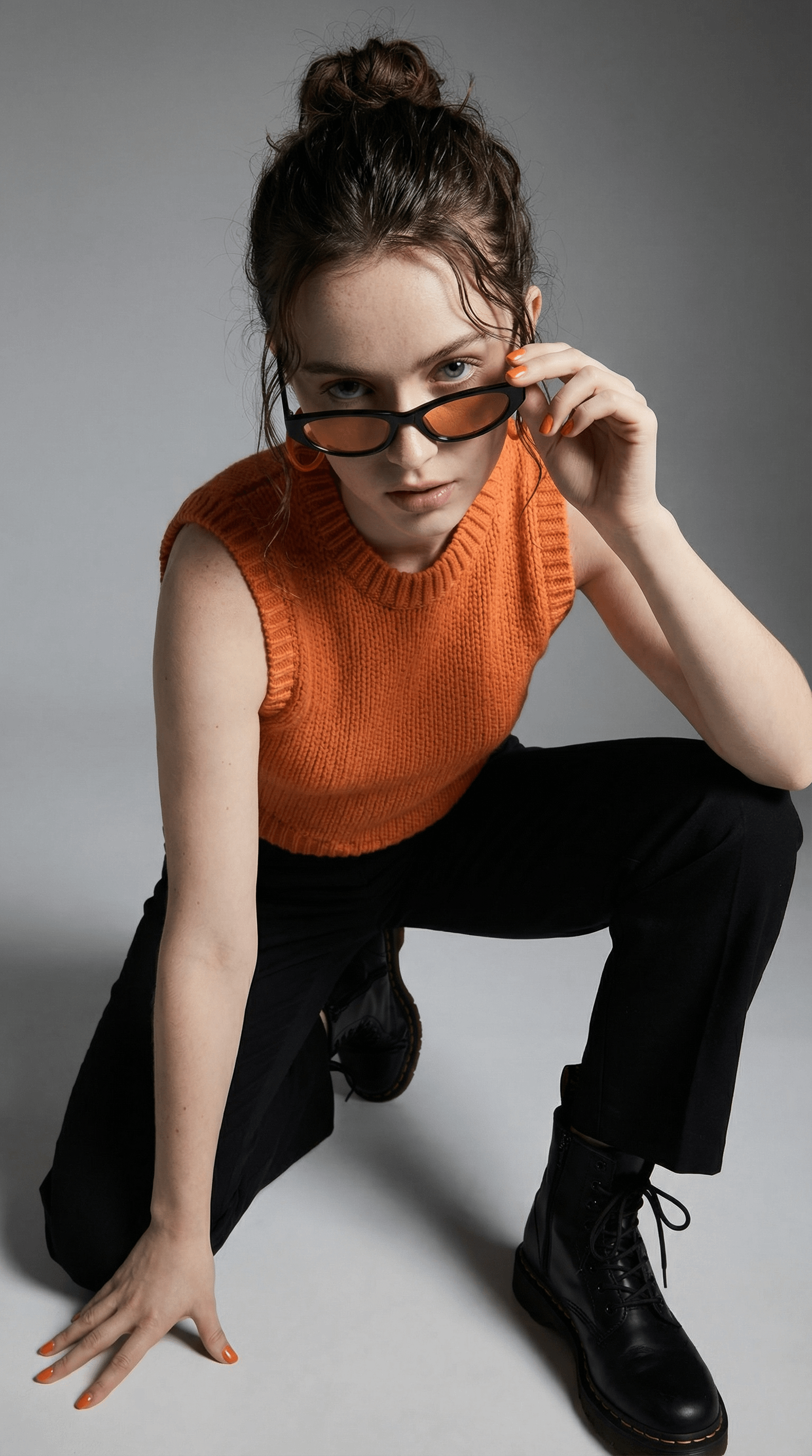 Close-up studio portrait of a woman wearing black sunglasses with orange tint, styled hair, and bright orange top against a light gray background.