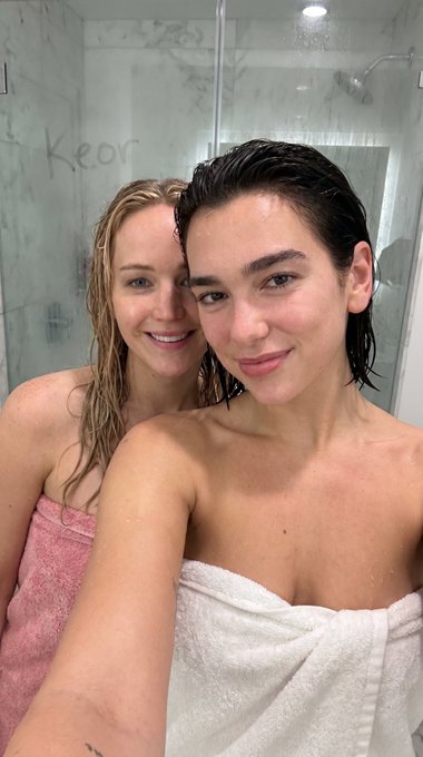 Selfie of two smiling individuals in a marble tiled shower, one in a pink towel and the other in a white towel, with water droplets on their skin.