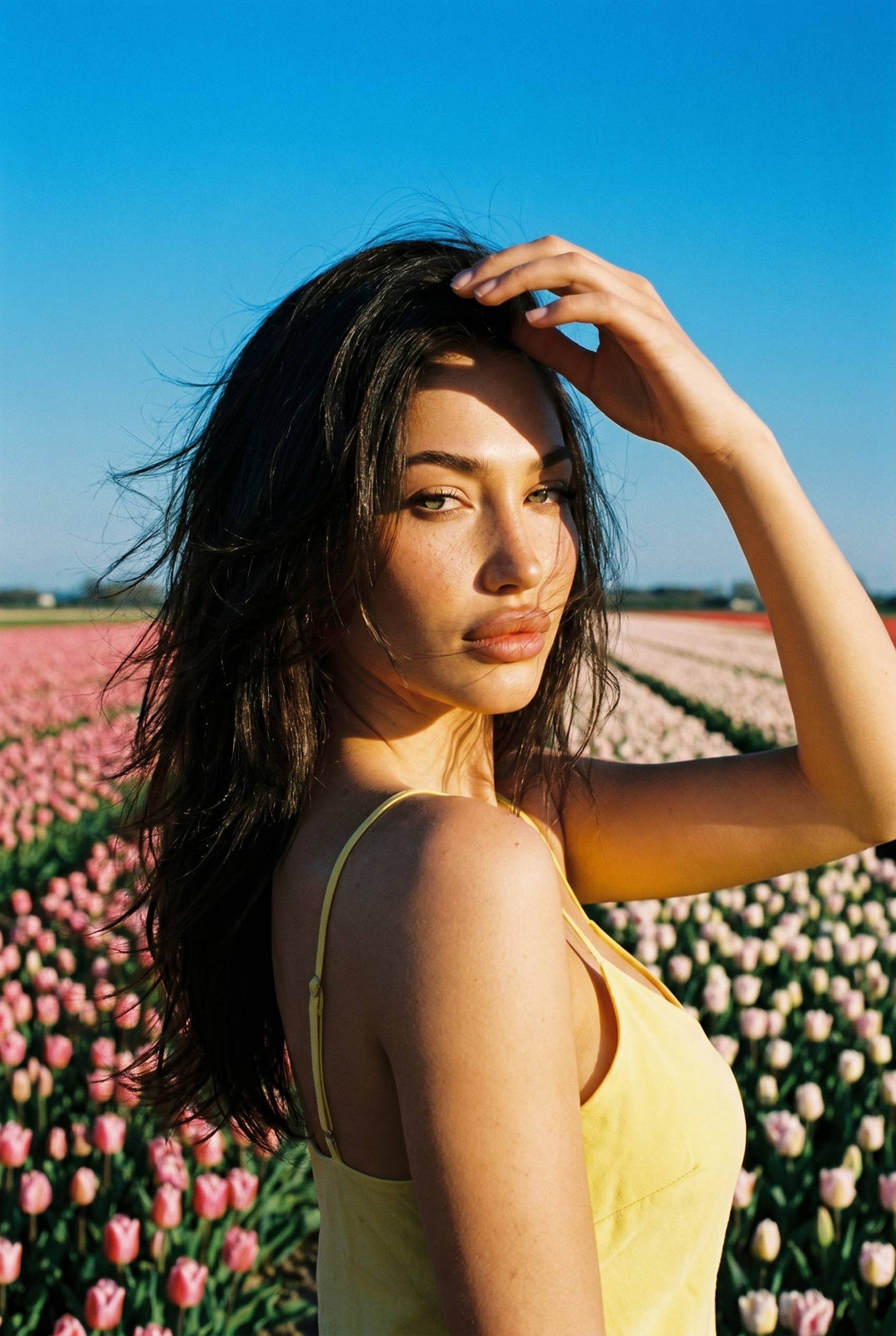 A woman in a yellow bikini standing in a vast tulip field, gazing back at the camera with a sultry expression.